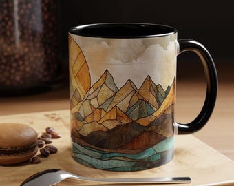 May include: A black ceramic mug featuring a colourful mountain scene design. The scene depicts a stylised mountain range with a sun rising over the peaks. The mug has a black handle.