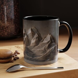 May include: A black ceramic mug with a mountain range design. The mountains are depicted in shades of gray and brown, with a textured appearance. The mug has a black handle.