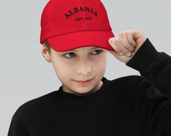 May include: A red baseball cap with the word "ALBANIA" arched across the front in black, with "EST. 1912" below. The cap is being held up by a person.