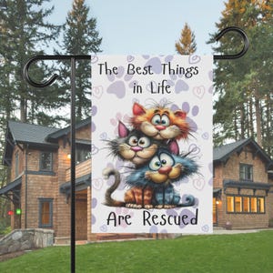 May include: A garden flag with the text "The Best Things in Life Are Rescued" and a cartoon image of three cats. The cats are orange, gray, and blue, with large eyes and expressive faces. The background has paw prints and hearts.