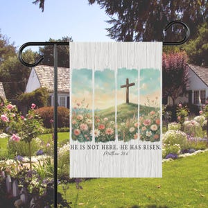 Peut inclure: Un drapeau de jardin avec une peinture à l'aquarelle d'une croix sur une colline avec des fleurs sauvages. Le drapeau porte le texte "HE IS NOT HERE. HE HAS RISEN. Matthew 28:6" sur un fond en bois blanc. Le drapeau est sur un support en métal noir.