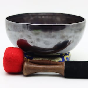 May include: A silver singing bowl with a red felt mallet and a wooden striker. The bowl is sitting on a blue and gold patterned cushion.