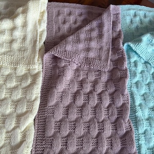 Peut inclure: Trois couvertures tricotées dans des couleurs pastel douces. Les couvertures sont de couleur crème, lavande et bleu clair. Chaque couverture présente un motif texturé en relief. Les couvertures sont pliées pour montrer les différentes couleurs et textures.