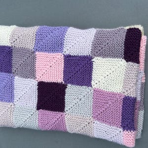 Puede incluir: Una manta de punto con un diseño de patchwork. La manta presenta cuadrados en tonos de morado, rosa y blanco. Los cuadrados están dispuestos en un patrón de cuadrícula, creando un textil visualmente atractivo y acogedor.
