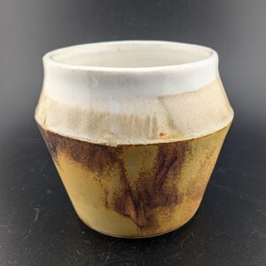 May include: A handmade ceramic cup with a unique design. The cup features a white rim, a tan middle section, and a dark brown base. The cup's exterior has a textured appearance, adding to its rustic charm. This piece is ideal for home decor or as a functional item.