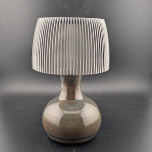 May include: A ceramic lamp with a brown and blue glazed base and a silver, vertically-lined shade. The lamp has a rounded base and a slender neck, with a glossy finish. The shade is cylindrical and made of vertical, parallel lines.