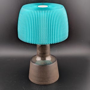May include: A teal-colored lamp with a ribbed lampshade and a dark brown base. The lampshade has a cylindrical shape with vertical ridges. The base is a dark brown color with a rounded top and a flared bottom. The lamp is on a black background.