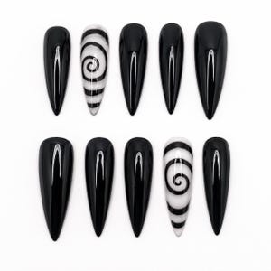 Op de afbeelding: Een set van tien stiletto-vormige press-on nagels met een glanzende afwerking. Acht nagels zijn effen zwart, terwijl twee nagels een zwart spiraalpatroon op een witte achtergrond hebben. De nagels zijn gerangschikt in twee rijen van vijf op wit.