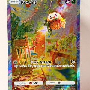 Op de afbeelding: Een holografische Rowlet Pokémon-kaart. De kaart toont een kleurrijke illustratie van Rowlet in een gedetailleerde binnenscène met meubels en planten. De kaart heeft de tekst "Fury Attack" en "HP 60".