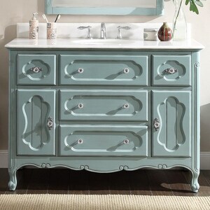 Puede incluir: Un mueble de ba&ntilde;o azul claro con encimera de m&aacute;rmol blanco. El mueble tiene m&uacute;ltiples cajones y puertas de armario con pomos de cristal. El dise&ntilde;o es ornamentado con detalles curvos y una base festoneada. Un espejo est&aacute; montado encima.