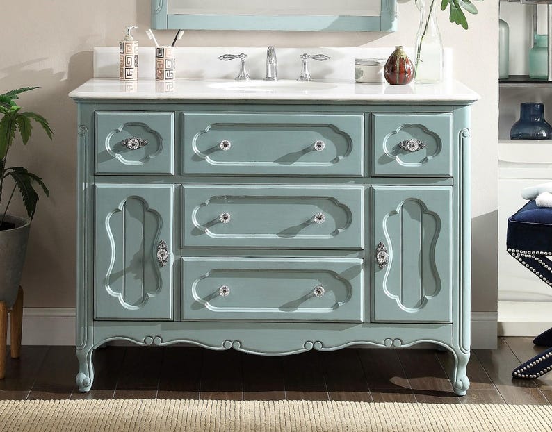Puede incluir: Un mueble de ba&ntilde;o azul claro con encimera de m&aacute;rmol blanco. El mueble tiene m&uacute;ltiples cajones y puertas de armario con pomos de cristal. El dise&ntilde;o es ornamentado con detalles curvos y una base festoneada. Un espejo est&aacute; montado encima.