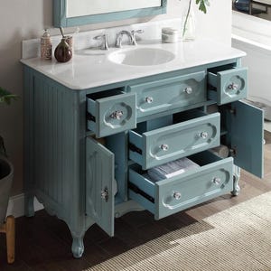 Puede incluir: Un mueble de ba&ntilde;o azul claro con encimera blanca y lavabo ovalado blanco. El mueble tiene m&uacute;ltiples cajones y puertas de armario con pomos de cristal. Los cajones est&aacute;n parcialmente abiertos, revelando espacio de almacenamiento. Un espejo es visible en el fondo.