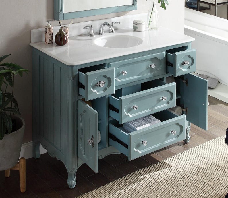 Puede incluir: Un mueble de ba&ntilde;o azul claro con encimera blanca y lavabo ovalado blanco. El mueble tiene m&uacute;ltiples cajones y puertas de armario con pomos de cristal. Los cajones est&aacute;n parcialmente abiertos, revelando espacio de almacenamiento. Un espejo es visible en el fondo.
