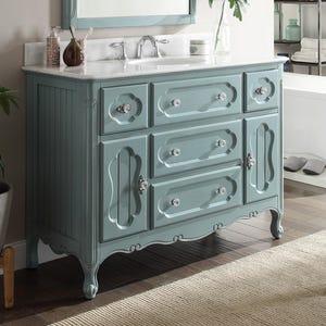 Puede incluir: Un mueble de ba&ntilde;o azul claro con encimera blanca y lavabo integrado. El mueble tiene varios cajones y puertas con pomos de cristal. La moldura decorativa y las patas curvas completan el dise&ntilde;o. Un espejo est&aacute; montado encima.