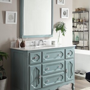 Puede incluir: Un mueble de ba&ntilde;o azul con un espejo enmarcado a juego. El mueble tiene m&uacute;ltiples cajones y puertas de armario con pomos de cristal. El espejo es rectangular con una parte superior decorativa. El mueble est&aacute; contra una pared neutra.