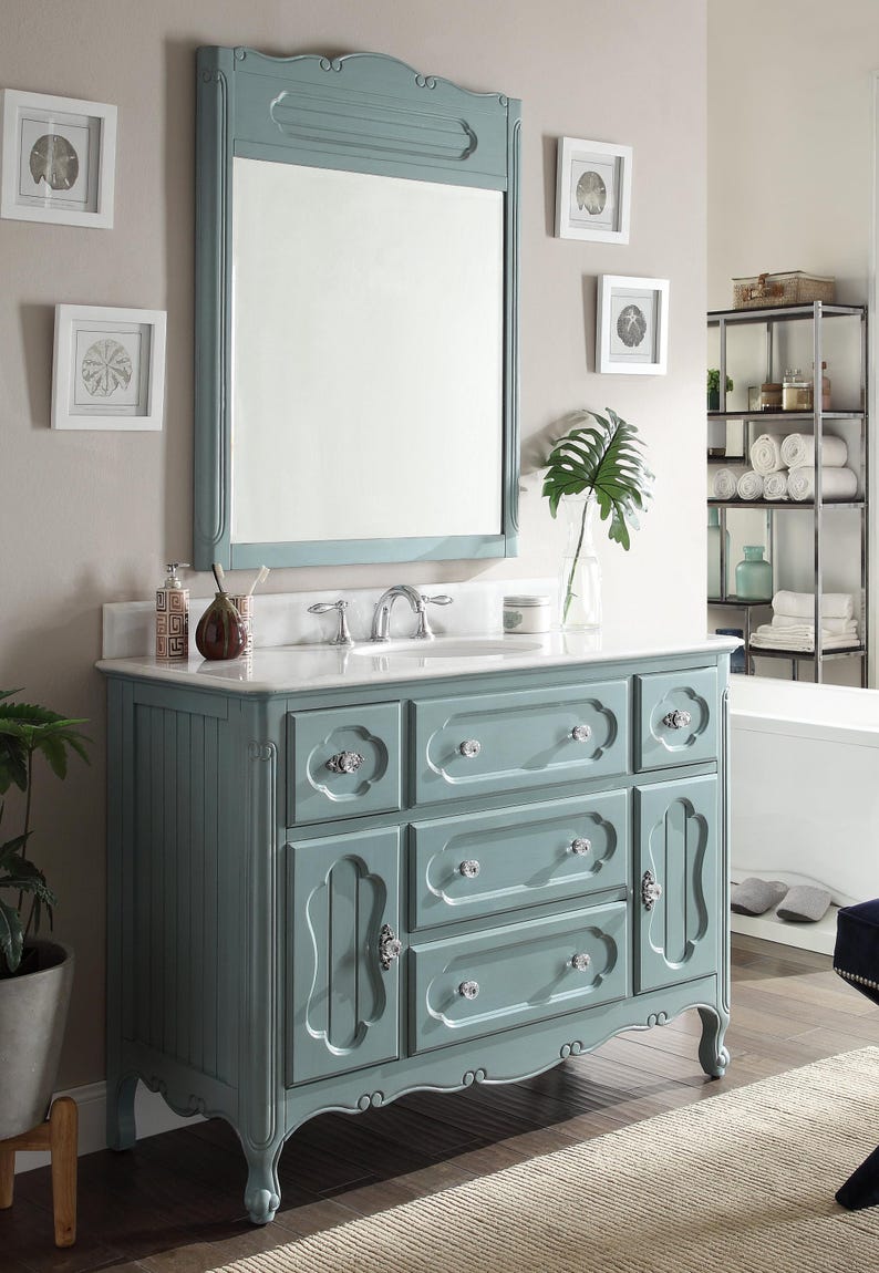 Puede incluir: Un mueble de ba&ntilde;o azul con un espejo enmarcado a juego. El mueble tiene m&uacute;ltiples cajones y puertas de armario con pomos de cristal. El espejo es rectangular con una parte superior decorativa. El mueble est&aacute; contra una pared neutra.