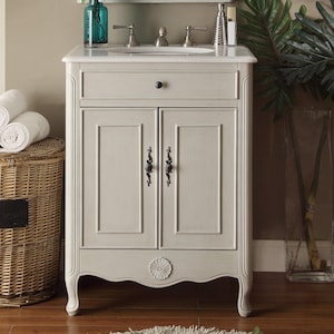 May include: A light gray bathroom vanity with a white countertop and a built-in sink. The vanity has two doors with decorative handles and a drawer. A wicker basket with rolled white towels sits to the left. A blue glass vase with flowers is on the countertop.
