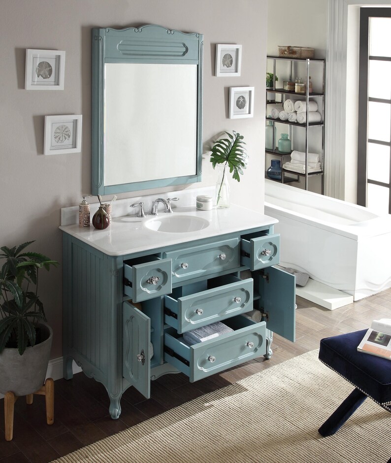 Puede incluir: Un mueble de ba&ntilde;o azul claro con encimera blanca y espejo enmarcado a juego. El mueble tiene m&uacute;ltiples cajones y puertas de armario con pomos de cristal. El ba&ntilde;o cuenta con un lavabo blanco y obras de arte enmarcadas.