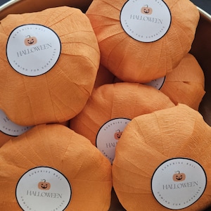 May include: A collection of orange crepe paper surprise balls shaped like pumpkins. Each ball features a circular white sticker with a jack-o'-lantern icon and the text HALLOWEEN SURPRISE BALL UNWIND FOR SURPRISES. The items are in a bowl.