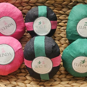 May include: Six colorful surprise balls in a woven basket. Two pink balls are labeled "Glinda", two black and green balls are labeled "W.C.E", and two green balls are labeled "Elphaba". Each ball is wrapped in crepe paper and has a circular label.