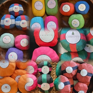 May include: A collection of round surprise balls wrapped in colorful crepe paper, each featuring a circular label with themed text and graphics including Easter, Valentine, Christmas, and Halloween designs, arranged in a group.