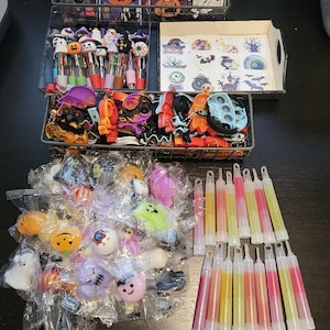 May include: A collection of Halloween-themed party favors including spiral-bound notebooks, multi-color ink pens, temporary tattoos, silicone fidget keychains, small plastic character figurines in clear bags, and various glow sticks in pink and yellow.