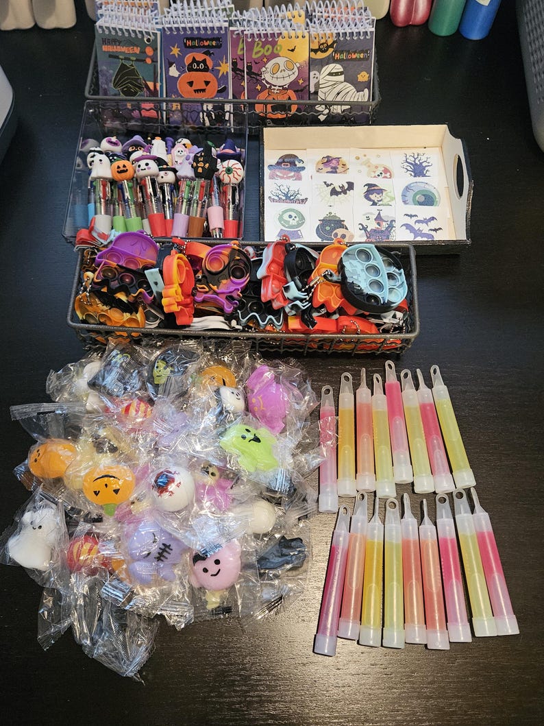 May include: A collection of Halloween-themed party favors including spiral-bound notebooks, multi-color ink pens, temporary tattoos, silicone fidget keychains, small plastic character figurines in clear bags, and various glow sticks in pink and yellow.