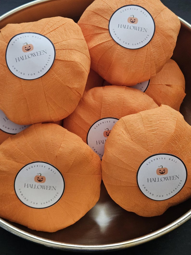 May include: A collection of orange crepe paper surprise balls shaped like pumpkins. Each ball features a circular white sticker with a jack-o'-lantern icon and the text HALLOWEEN SURPRISE BALL UNWIND FOR SURPRISES. The items are in a bowl.