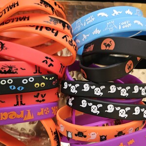 May include: A collection of silicone wristbands in orange, black, purple, and light blue. The bands feature various Halloween-themed graphics including ghosts, bats, cats, eyes, and candy, with text such as "Happy Halloween" and "Trick or Treat."