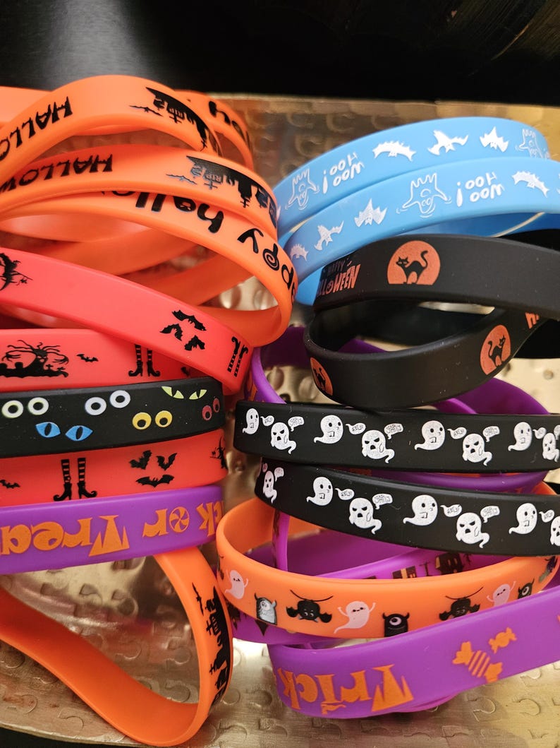 May include: A collection of silicone wristbands in orange, black, purple, and light blue. The bands feature various Halloween-themed graphics including ghosts, bats, cats, eyes, and candy, with text such as "Happy Halloween" and "Trick or Treat."