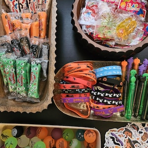 May include: A collection of Halloween party favors including Play-Doh containers, sticky toys, silicone wristbands with text like Trick or Treat and Halloween, bubble wands with skull toppers, colorful bouncy balls, and assorted holiday-themed stickers.