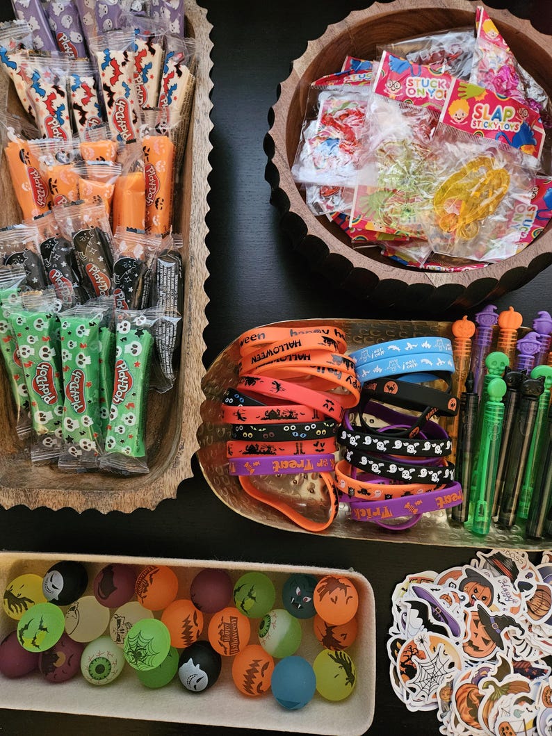 May include: A collection of Halloween party favors including Play-Doh containers, sticky toys, silicone wristbands with text like Trick or Treat and Halloween, bubble wands with skull toppers, colorful bouncy balls, and assorted holiday-themed stickers.