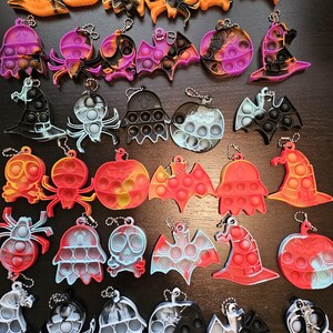 May include: A collection of silicone sensory fidget keychains in various Halloween-themed shapes including pumpkins, ghosts, bats, spiders, skulls, and witch hats. Each keychain features a push-pop bubble design and a metal ball chain attachment.