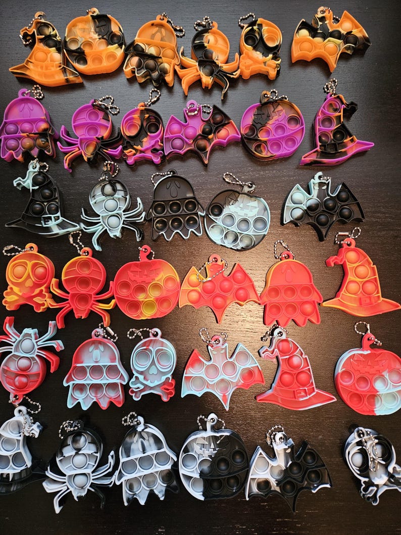 May include: A collection of silicone sensory fidget keychains in various Halloween-themed shapes including pumpkins, ghosts, bats, spiders, skulls, and witch hats. Each keychain features a push-pop bubble design and a metal ball chain attachment.