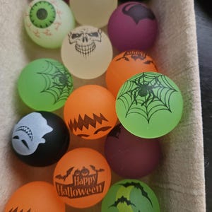 May include: A collection of round rubber bouncy balls featuring various Halloween-themed designs including skulls, spiders, spiderwebs, eyeballs, ghosts, bats, and pumpkins in orange, green, purple, black, and off-white colors.