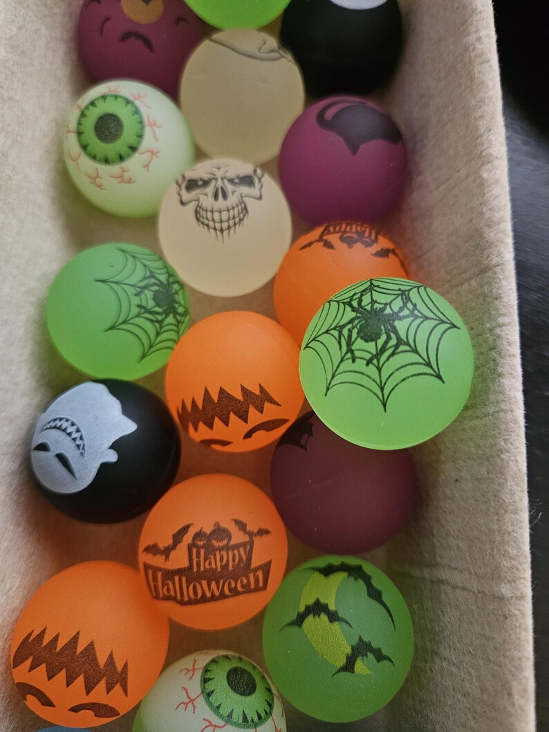 May include: A collection of round rubber bouncy balls featuring various Halloween-themed designs including skulls, spiders, spiderwebs, eyeballs, ghosts, bats, and pumpkins in orange, green, purple, black, and off-white colors.