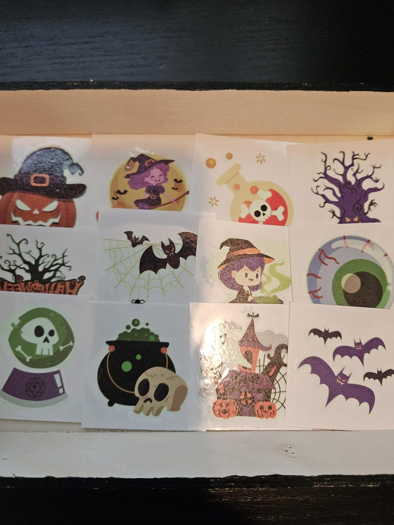 May include: A collection of twelve square temporary tattoos featuring Halloween themes including a jack-o'-lantern, a witch, a potion bottle, a skull, a haunted house, bats, a spider web, a green eyeball, and a crystal ball with a skull.