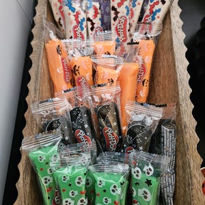 May include: A collection of small, individually wrapped Play-Doh sticks arranged in a wooden tray. The wrappers feature various Halloween-themed patterns, including white with bats, orange with jack-o'-lanterns, black with cats, and green with skulls.