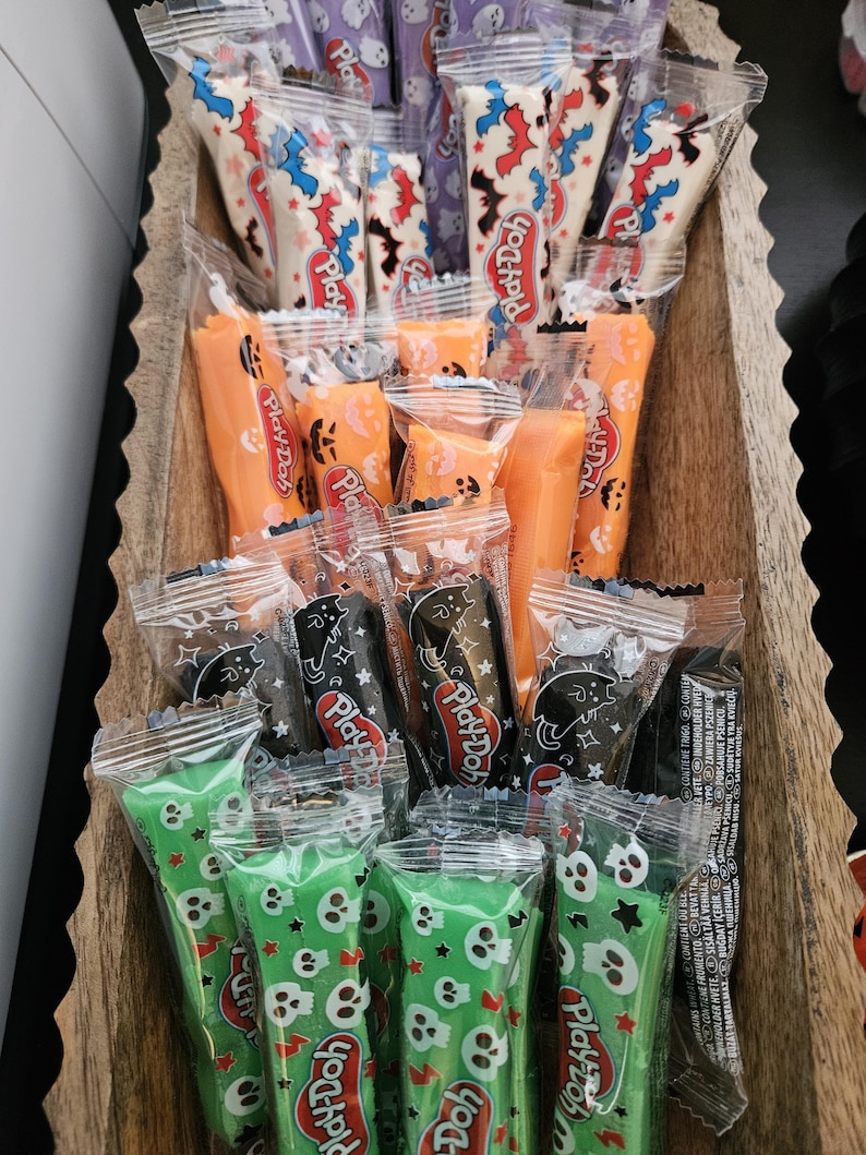 May include: A collection of small, individually wrapped Play-Doh sticks arranged in a wooden tray. The wrappers feature various Halloween-themed patterns, including white with bats, orange with jack-o'-lanterns, black with cats, and green with skulls.