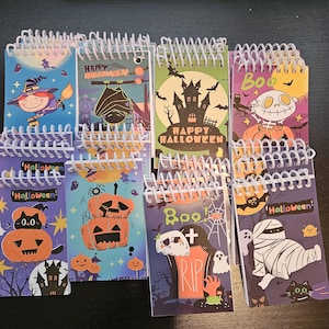 May include: A collection of small spiral-bound notebooks featuring various Halloween-themed illustrations including pumpkins, ghosts, a mummy, a witch, a bat, and a haunted house, with text such as Happy Halloween and Boo printed on the covers.