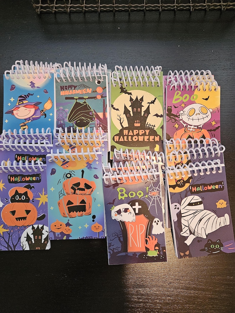 May include: A collection of small spiral-bound notebooks featuring various Halloween-themed illustrations including pumpkins, ghosts, a mummy, a witch, a bat, and a haunted house, with text such as Happy Halloween and Boo printed on the covers.