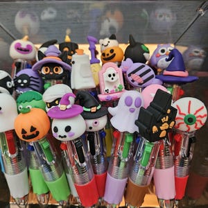 May include: A collection of multi-color ink pens featuring various Halloween-themed toppers including pumpkins, ghosts, cats in witch hats, skulls, eyeballs, and haunted houses. Each pen has a clear barrel with multiple colored ink sliders.