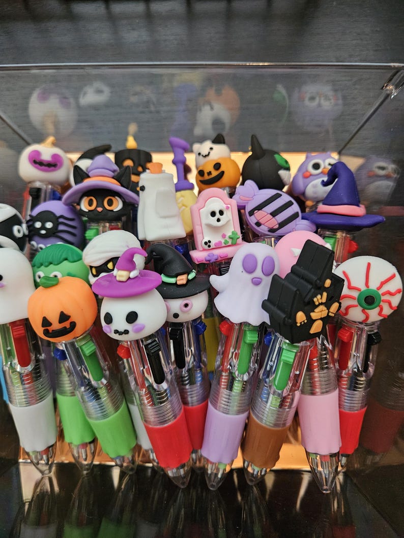 May include: A collection of multi-color ink pens featuring various Halloween-themed toppers including pumpkins, ghosts, cats in witch hats, skulls, eyeballs, and haunted houses. Each pen has a clear barrel with multiple colored ink sliders.