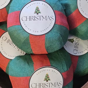 May include: A collection of Christmas surprise balls. Each ball is wrapped in teal and green crepe paper with a red band. A white circular label features a Christmas tree graphic and the word "CHRISTMAS". The text "SURPRISE BALL" and "UNWIND FOR SURPRISES" are also on the label.
