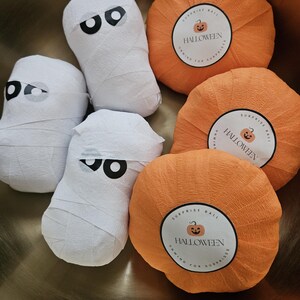 May include: Three white mummy-shaped figures wrapped in paper with black and white eye decals, and three orange pumpkin-shaped paper balls. Each pumpkin features a circular label with a jack-o'-lantern icon and the text HALLOWEEN SURPRISE BALL.