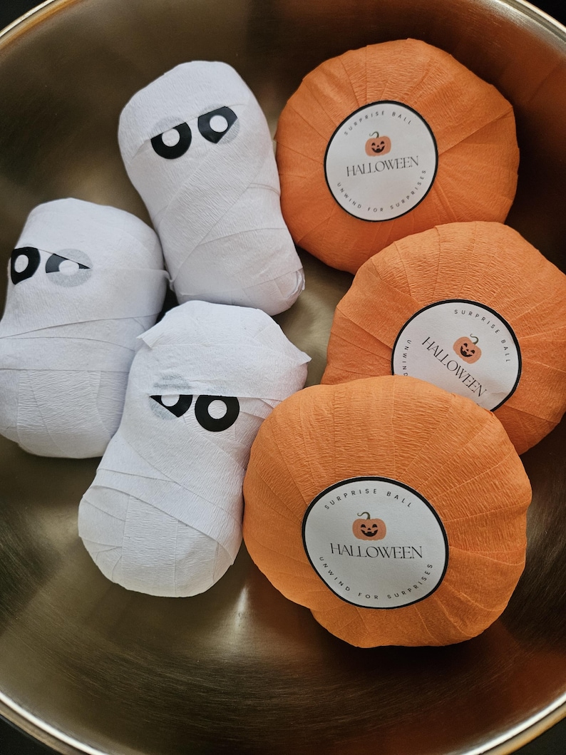 May include: Three white mummy-shaped figures wrapped in paper with black and white eye decals, and three orange pumpkin-shaped paper balls. Each pumpkin features a circular label with a jack-o'-lantern icon and the text HALLOWEEN SURPRISE BALL.