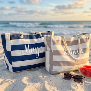 May include: Two large tote bags with rope handles. One bag features navy blue and white horizontal stripes with the name Maya in white script. The other bag has beige and white horizontal stripes with the name Isabelle in white script.