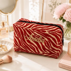 K&ouml;nnte beinhalten: Eine rechteckige Rei&szlig;verschlusstasche mit einem rot-beigen Zebramuster. Auf der Vorderseite ist der Name Amelia in einer goldfarbenen Schreibschrift eingestickt. Die Tasche hat einen schwarzen Rei&szlig;verschluss an der Oberkante.