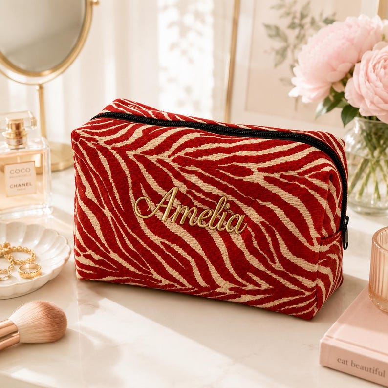 K&ouml;nnte beinhalten: Eine rechteckige Rei&szlig;verschlusstasche mit einem rot-beigen Zebramuster. Auf der Vorderseite ist der Name Amelia in einer goldfarbenen Schreibschrift eingestickt. Die Tasche hat einen schwarzen Rei&szlig;verschluss an der Oberkante.