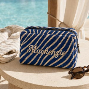 K&ouml;nnte beinhalten: Eine rechteckige Rei&szlig;verschlusstasche mit einem diagonalen Streifenmuster in Marineblau und Beige. Der Name Mackenzie ist in beiger Schreibschrift mittig auf der Vorderseite des strukturierten Stoffes eingestickt. Schwarzer Rei&szlig;verschluss.